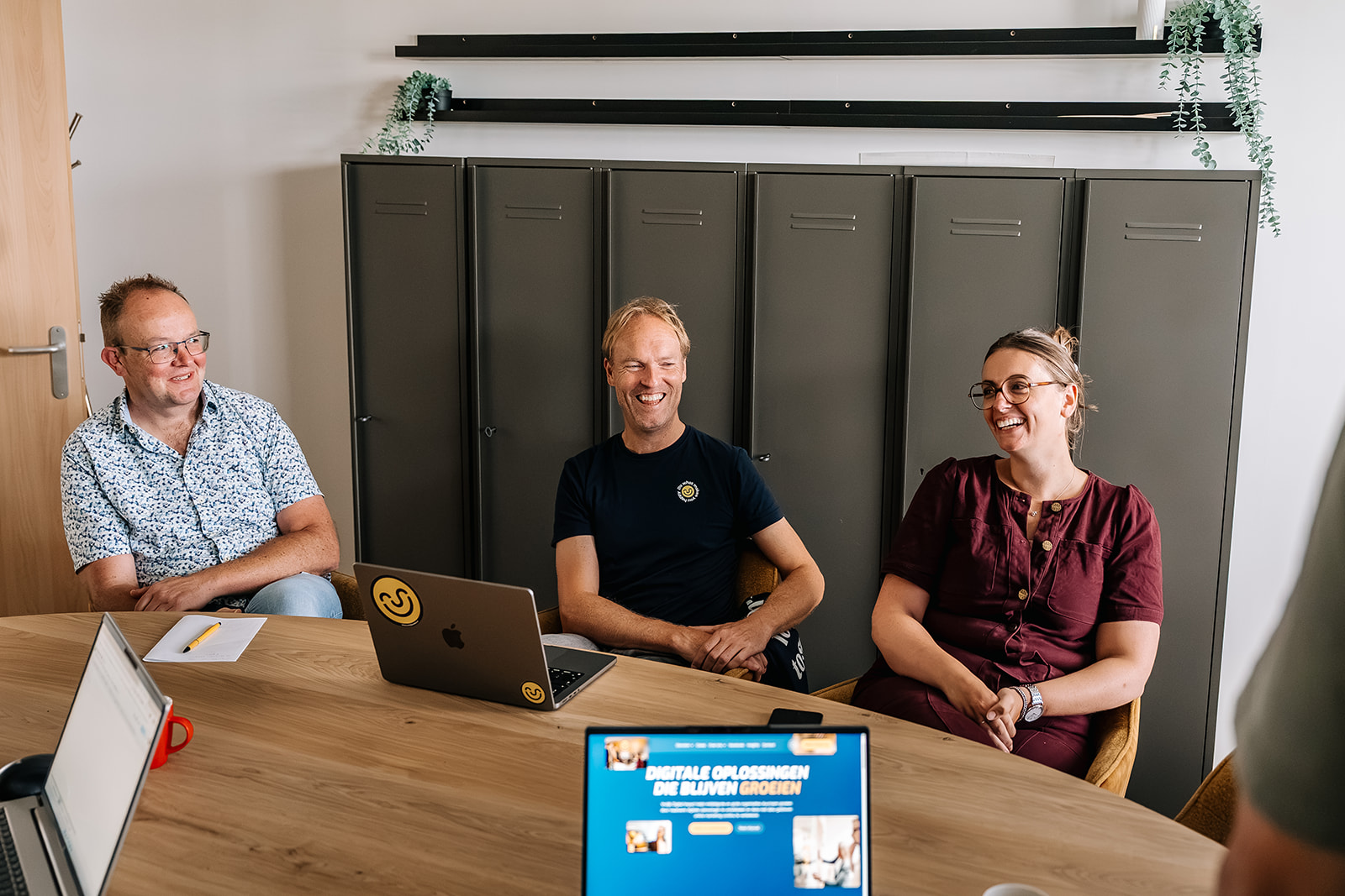 Drie teamleden zitten lachend achter een houten bureau met laptops in een modern kantoor met grijze kasten
