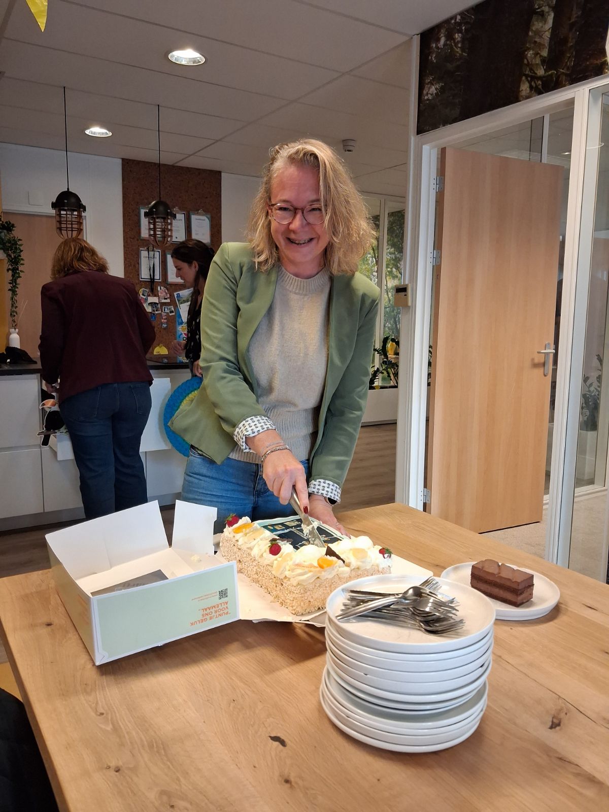 Vrouw met groen jasje snijd taart aan en lacht vrolijk