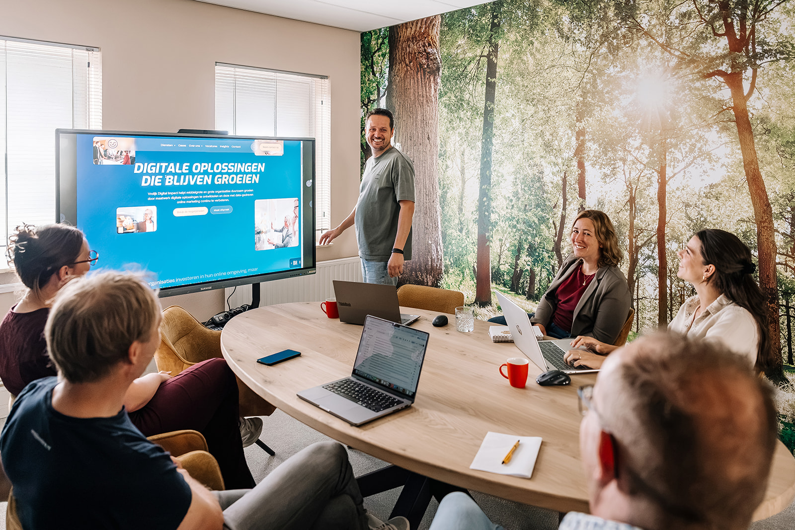 Team presenteert digitale oplossingen op scherm tijdens kantoorvergadering met laptops en forest wallpaper