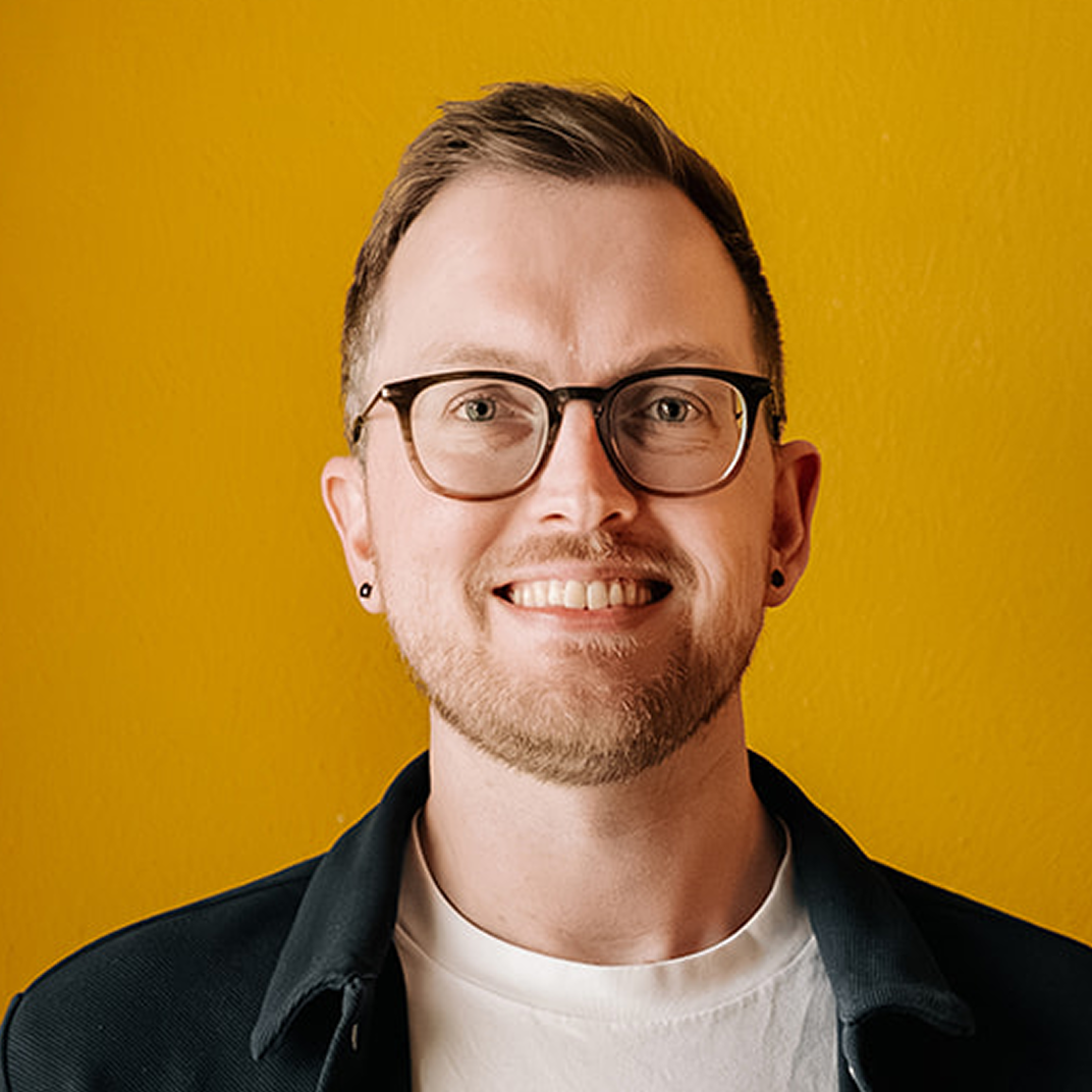 man met bril wit shirt en jasje lachend voor een gele muur
